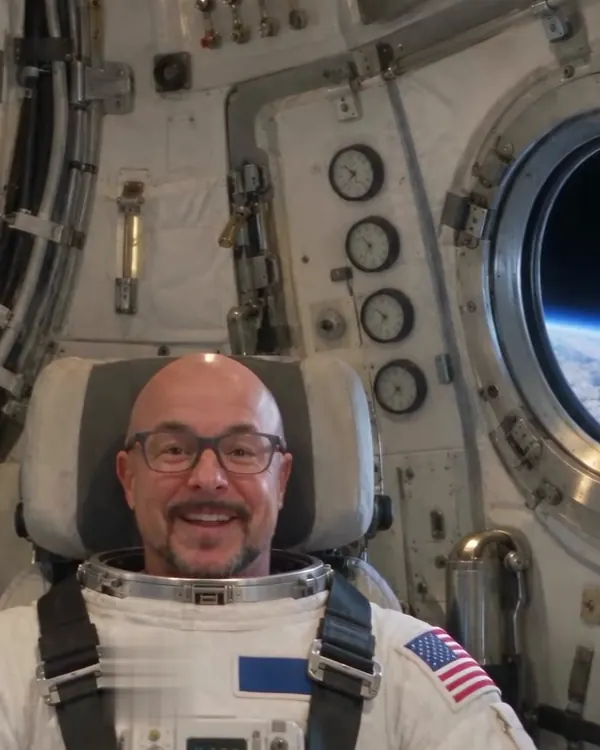 The same presenter in a spacesuit, strapped into a spacecraft seat with Earth through the porthole